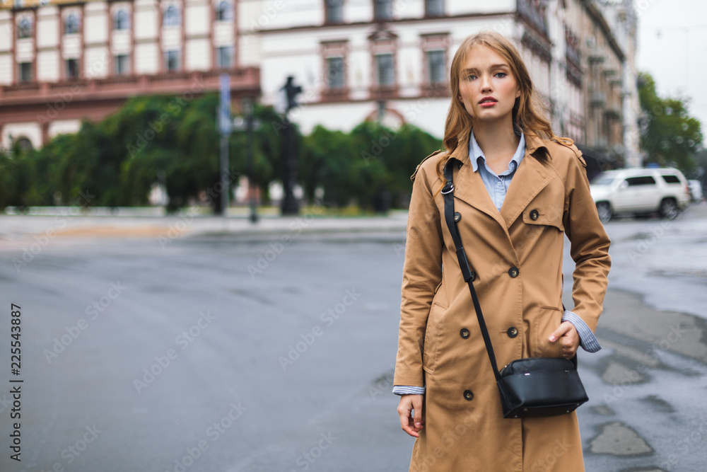 Beautiful pensive girl in trench coat with little black cross bag dreamily looking in camera while spending time on cozy city street