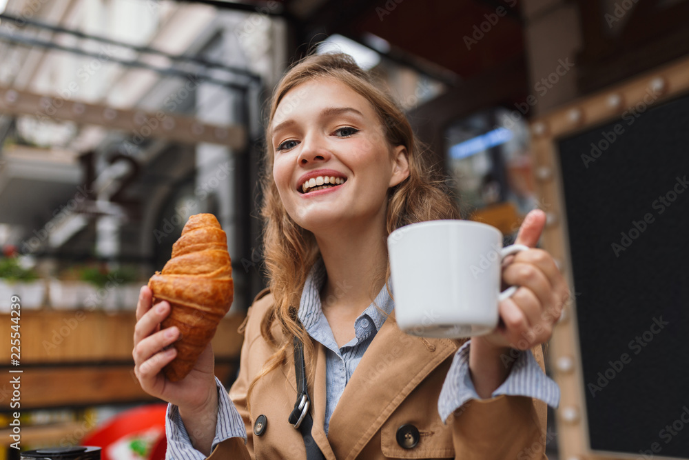 Portrait of beautiful smiling girl in trench coat happily looking in camera holding croissant and white cup in hands while spending time outdoor at cozy cafe terrace