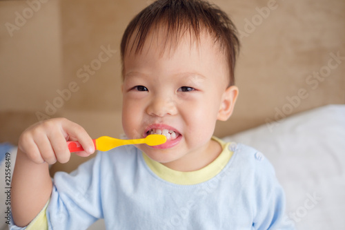 Cute smiling little Asian 30 months / 2 year old toddler boy child wearing pajama sitting in bed holding toothbrush and learn to brushing teeth in the morning at home, Tooth care for children concept