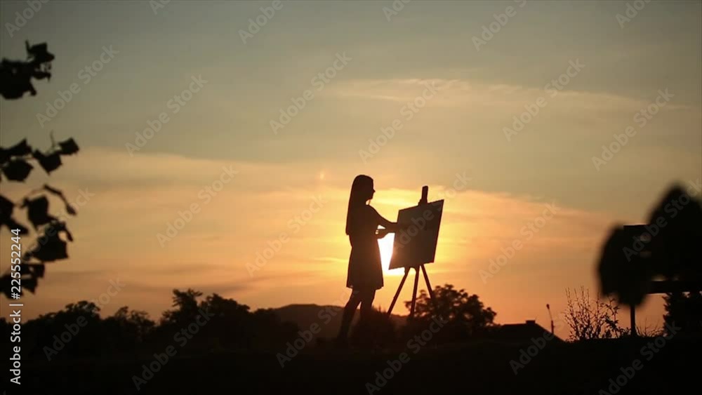 Silhouette of a blonde girl paints a painting on the canvas with the help of paints. A wooden easel keeps the picture. Summer is a sunny day, sunset