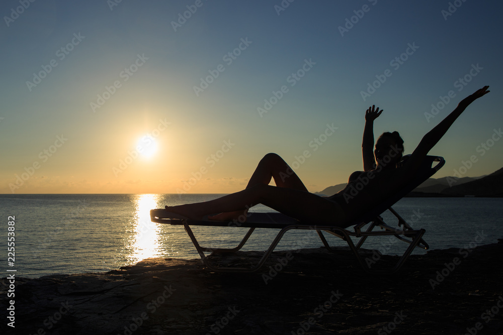 Silhouette of the young woman is lying on the deckchair at sunset.