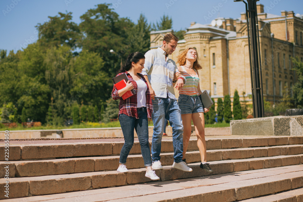 Obraz premium Happy students are walking together on campus. One man with two girls. Students between lectures getting sown the stairs in a park