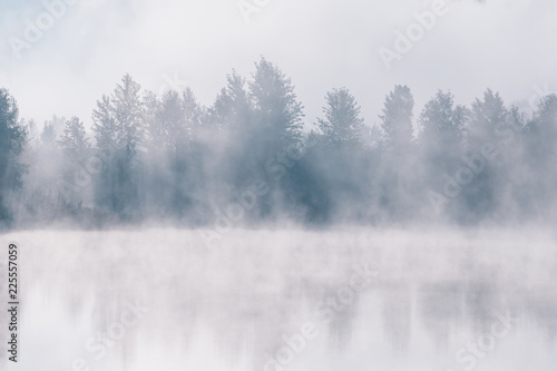 Foggy river coast at the autumn morning. Reflections of forest trees in the water.