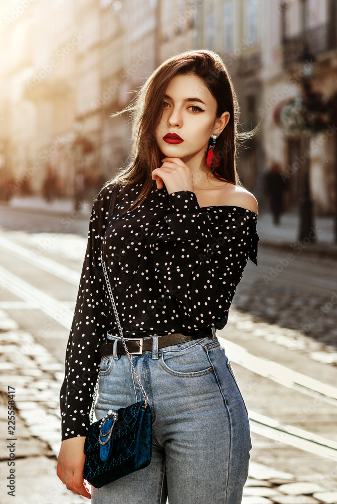 Outdoor portrait of yong beautiful fashionable woman wearing stylish black  white polka dot blouse, blue high waisted mom jeans, with small quilted  bag. Model posing in street of european city Stock Photo
