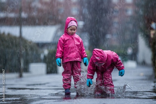 Girls are having fun in water on street in cold autumn day, girls splashing water in rain, happy and cheerful girls enjoying cold weather, kids in pink rain coats and rubber boots  © Ji