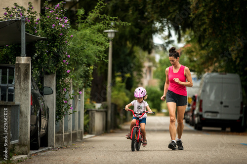 Beautiful and happy young mother teaching her cute daughter to ride a bicycle. Both smiling, Summer street in background, child in pink helmet enjoying first bike ride, healthy and sporty family