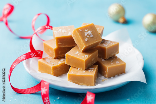 Caramel fudge candies on a plate with Christmas red ribbon on blue background. Close up. photo