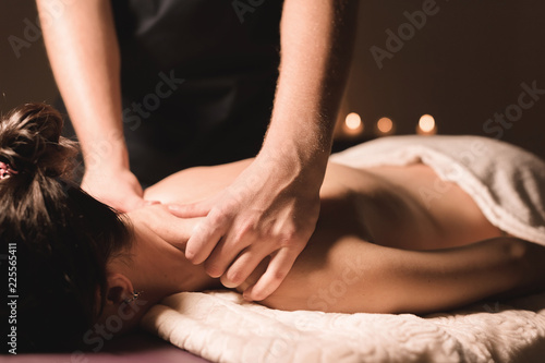 Men s hands make a therapeutic neck massage for a girl lying on a massage couch in a massage spa with dark lighting. Close-up. Dark Key
