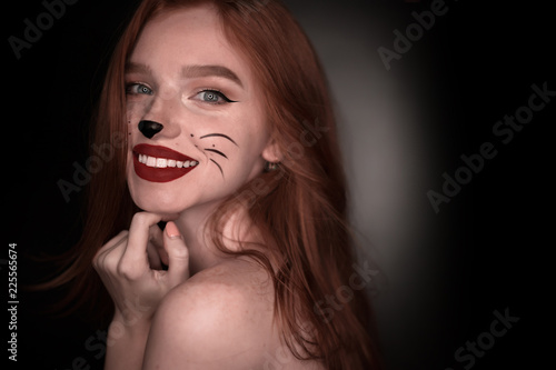 Cheerful beautiful redhead girl woman with cat carnival halloween makeup posing isolated over black wall background looking camera near empty copyspace.