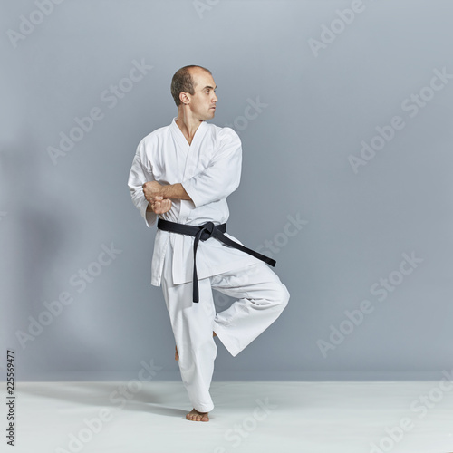Sportsman in a white karategi and with a black belt trains formal karate exercises on a gray background