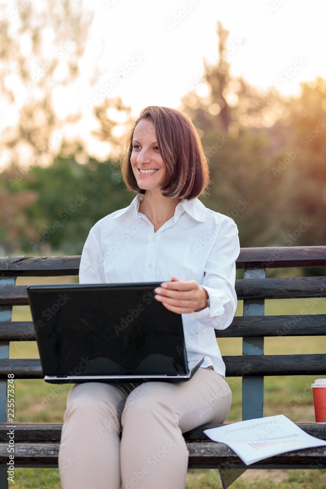 Smiling young businesswoman working on a laptop in a city park, sitting on a bench. Work anywhere concept. Smiling young businesswoman working on a laptop in a city park, sitting on a bench. Work anywhere concept.