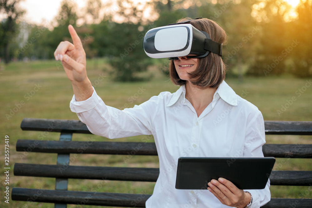 Fototapeta premium Young businesswoman using virtual reality goggles while sitting on a bench in a city park and working.