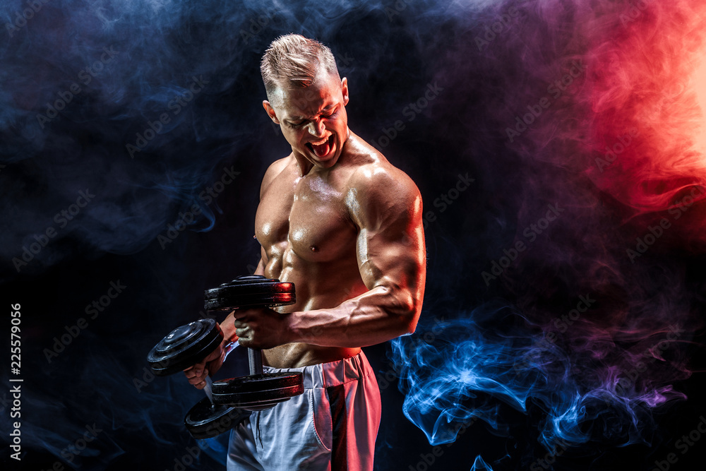 Topless man exercising biceps with dumbbells posing in studio fuul of colored smoke