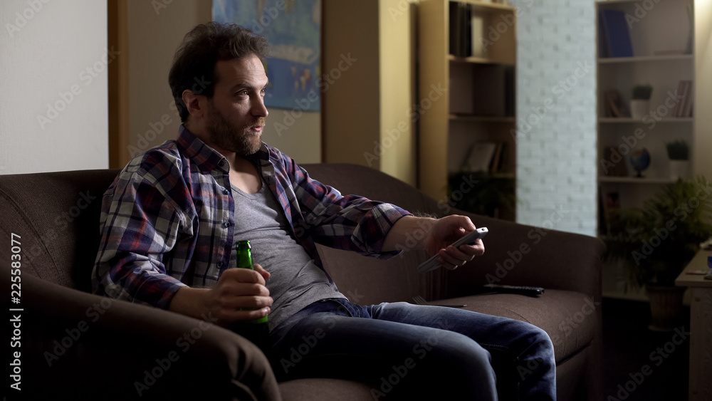Guy sitting on sofa with beer bottle in hand, using remote control to switch TV