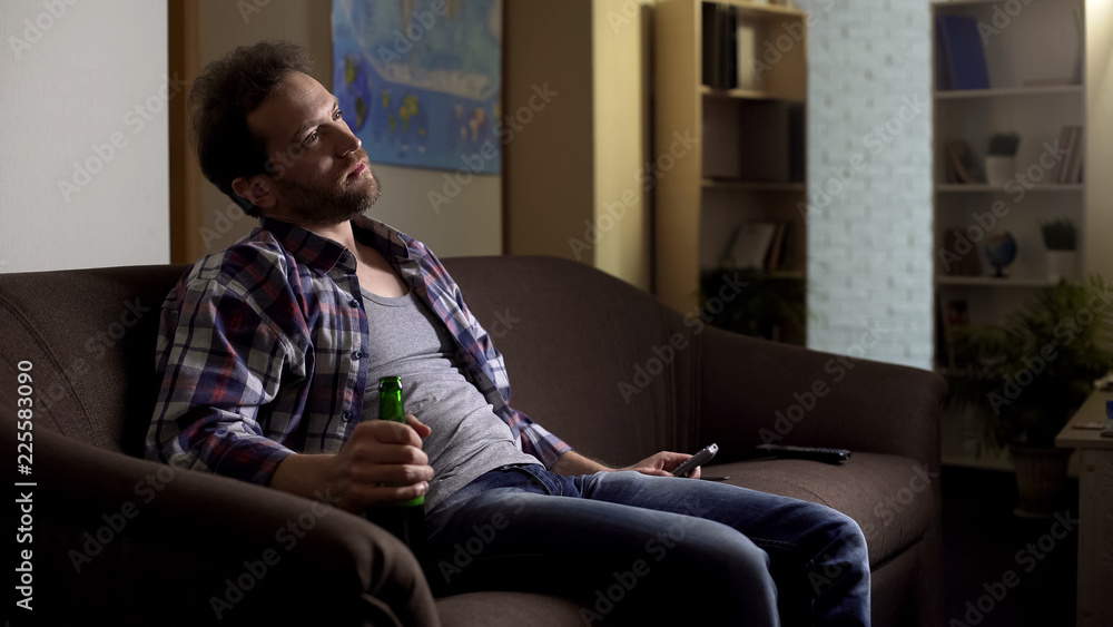 Grubby man sitting on sofa with beer bottle in hand and watching TV, alcoholism