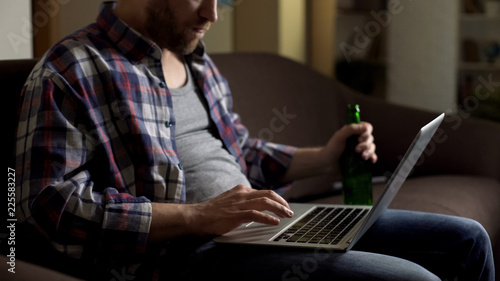 Untidy man on sofa using laptop and drinking beer, browsing internet, loneliness