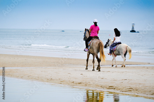 Back of young asian girls ridding horse on the beach with lighthouse in far background in the sea.