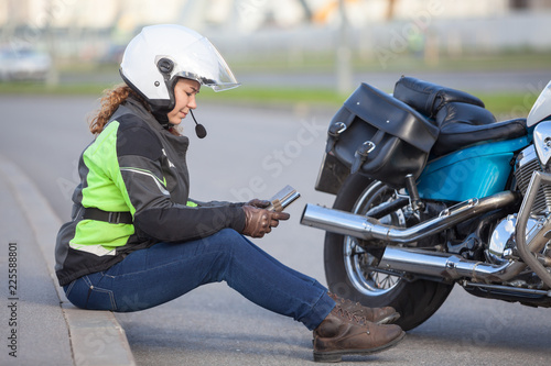Motorcyclist woman typing a message in her smartphone while sitting on roadside near motorcycle