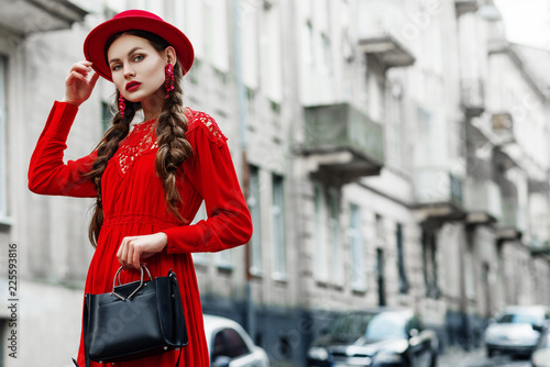 Foto Outdoor portrait of young beautiful fashionable woman wearing total red look: dress, hat, earrings, holding black leather handbad, bag, posing in street of european city