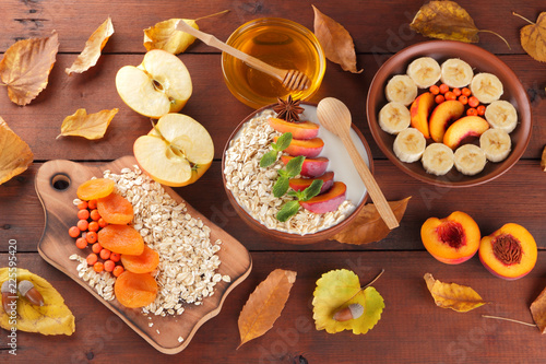 Oatmeal with different fruits and honey. Oat flakes with milk and dried apricots on wooden background. Healthy food for vegan. Dry oats with berries of mountain ash and green mint. Autumn pattern