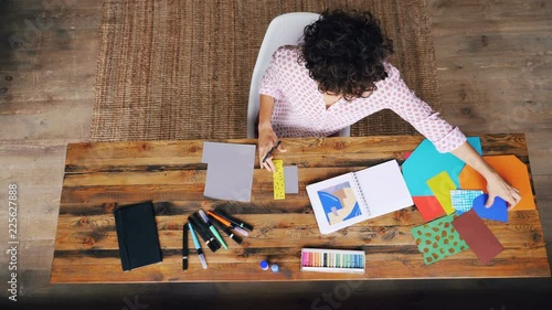 Top view of curly-headed woman making colorful paper collage sitting at table cutting figures with scissors. Design, creative people and interiors concept.