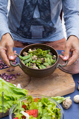 Woman hands hold a copper pot with cooked green beans, broccoli and mushrooms vegetables. Vegan vegetarian healthy food. Lunch or dinner.