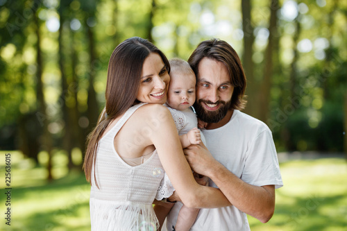 Adorable mom, handsome father and their wonderful little daughter dressed in the white just clothes are embracing in the park.