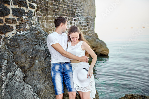 Couple in love hugging on the rocks against the background of an old stone wall and sea water. Young man and attractive woman in a white summer dress with a hat in her hand. Engagement portrait