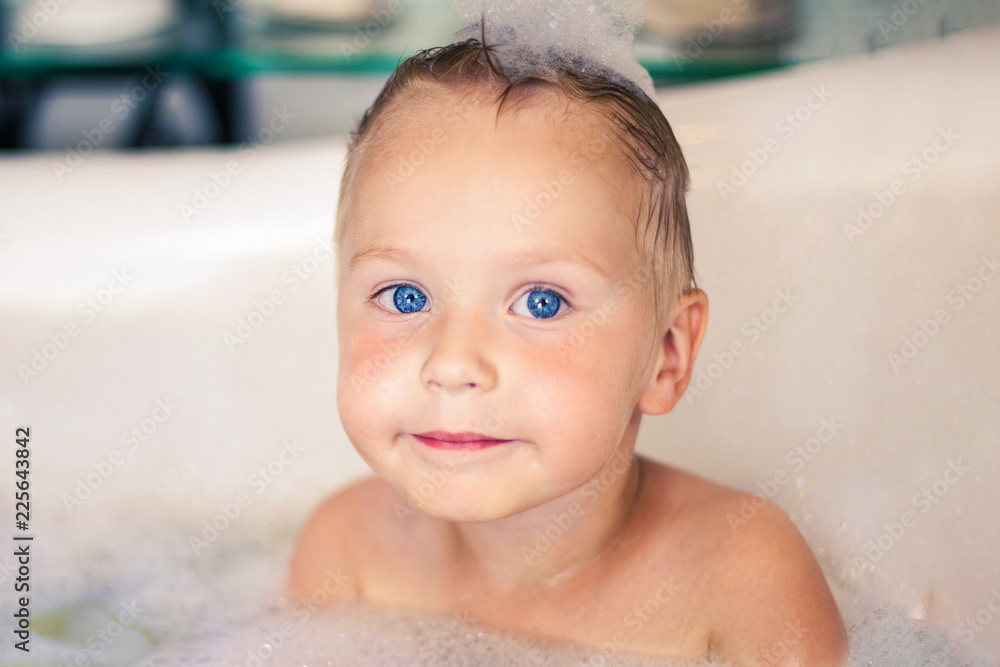 Cute caucasian blonde boy with bright blue eyes sits in the bathtube, with foam bubbles on the head. Expressive look. Indoors, copy space. Cute caucasian blonde boy with bright blue eyes sits in the bathtube, with foam bubbles on the head. Expressive look. Indoors, copy space.