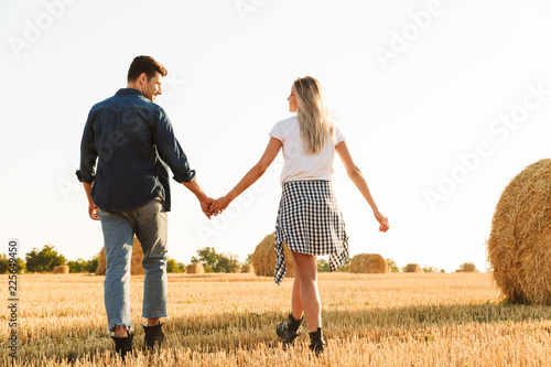 Photo of amazing couple man and woman walking through golden field with bunch of haystacks, during sunny day