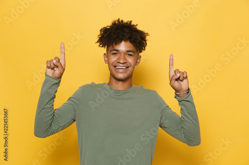 Portrait of joyous african guy having stylish afro hairdo pointing fingers upward at copyspace, isolated over yellow background photo