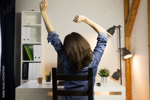 Woman stretching her arms sitting by the table at work next to computer. Working late concept photo