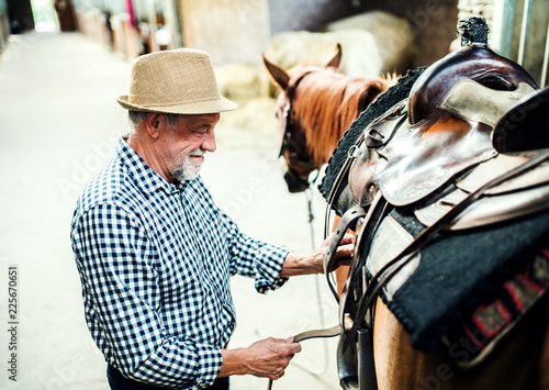 A senior man putting a saddle on a horse in a stable.