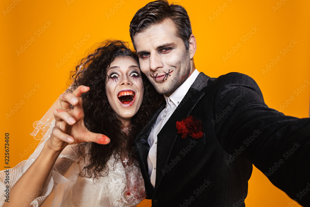 Photo of spooky zombie couple bridegroom and bride wearing wedding outfit and halloween makeup laughing while taking selfie, isolated over yellow background Photo of spooky zombie couple bridegroom and bride wearing wedding outfit and halloween makeup laughing while taking selfie, isolated over yellow background
