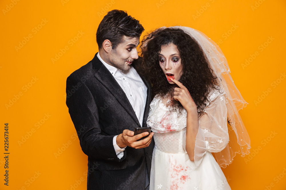 Photo of excited zombie couple bridegroom and bride wearing wedding outfit and halloween makeup using smartphone, isolated over yellow background Photo of excited zombie couple bridegroom and bride wearing wedding outfit and halloween makeup using smartphone, isolated over yellow background