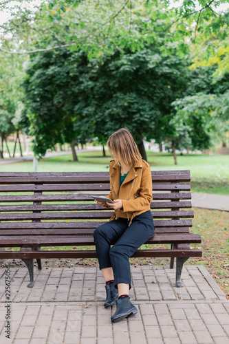 Beautiful girl is sitting on a bench with a tablet in the park.