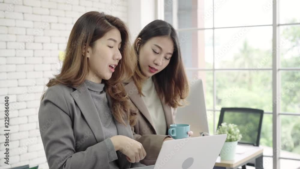 Two Asian young creative businesswomen holding a cup of coffee, working on laptop and disscuss about work while working in office. Casual business, freelance work at cafe, social meeting concept. Two Asian young creative businesswomen holding a cup of coffee, working on laptop and disscuss about work while working in office. Casual business, freelance work at cafe, social meeting concept.