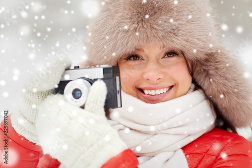 people, technology and leisure concept - happy woman in winter fur hat taking picture by film camera outdoors