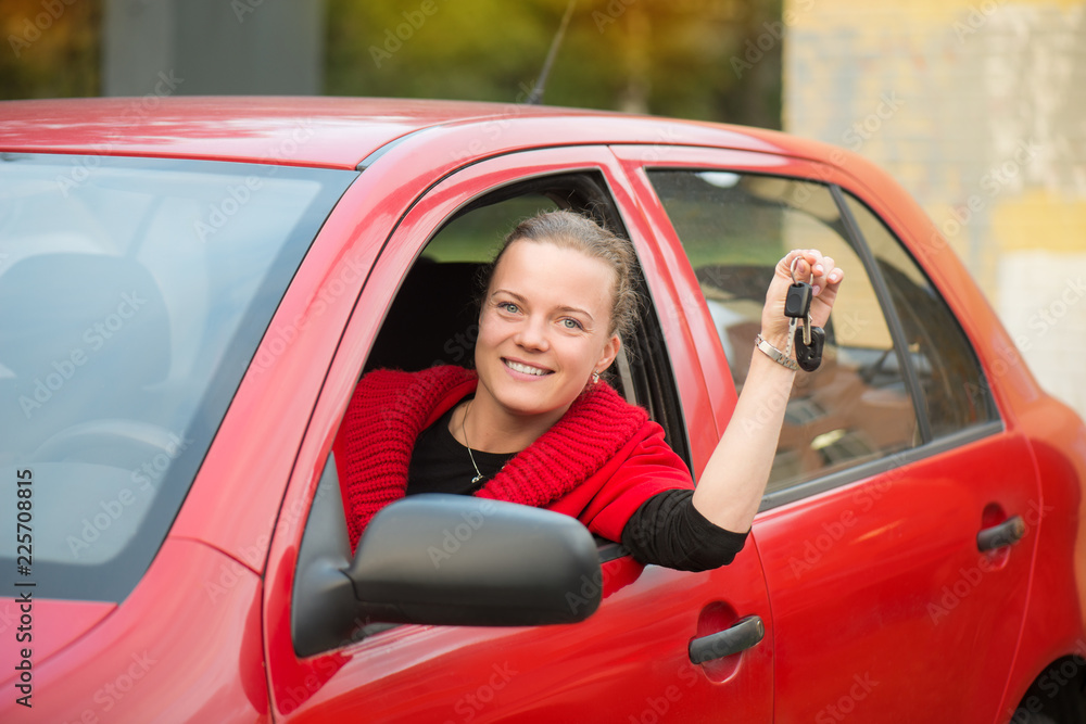 A beautiful young girl dressed in a red coat sits behind the wheel of a red car, smiles and holds car keys