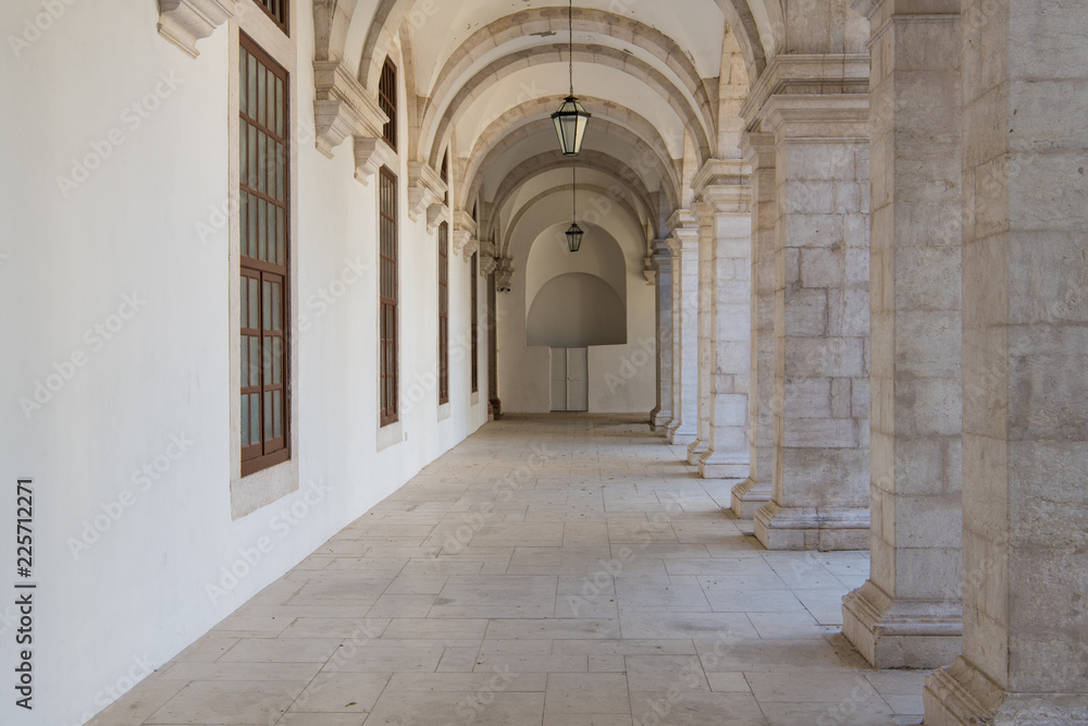 Long passageway moving to perspective under arches with stone block columns on one side and a white wall on the other side in the cloisters at the Convento da Graca in Lisbon, Portugal