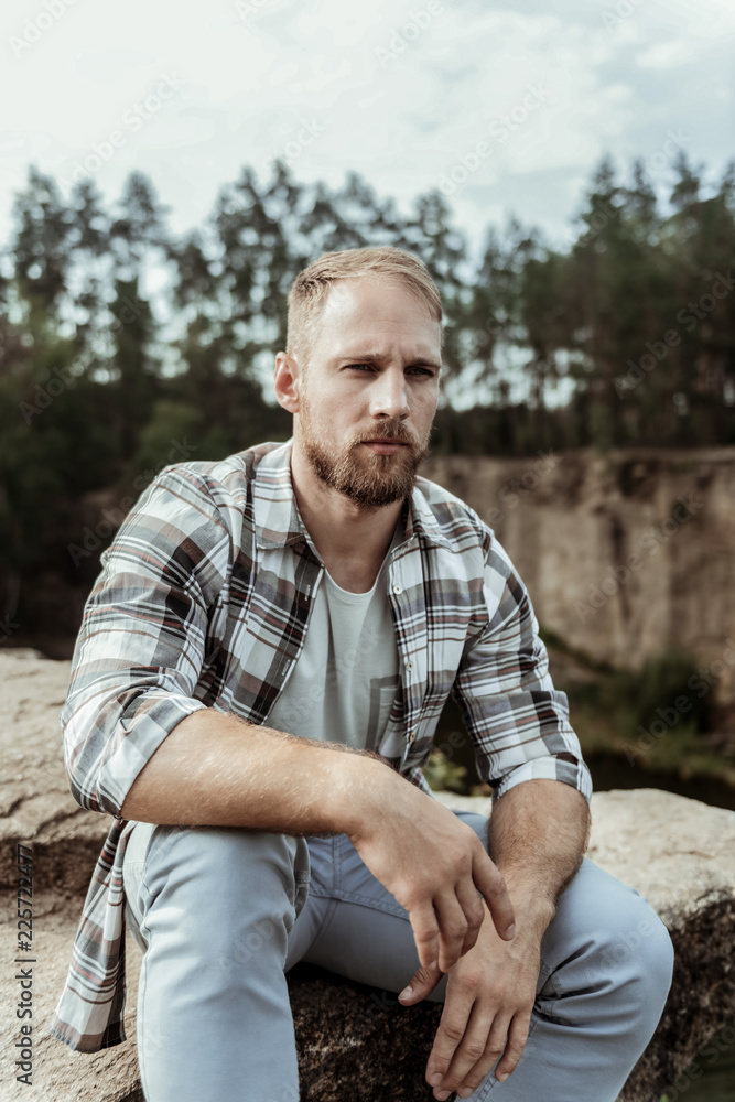 Blonde-haired model. Handsome blonde-haired model wearing jeans and squared shirt posing for photo while sitting on rock near canyon Blonde-haired model. Handsome blonde-haired model wearing jeans and squared shirt posing for photo while sitting on rock near canyon