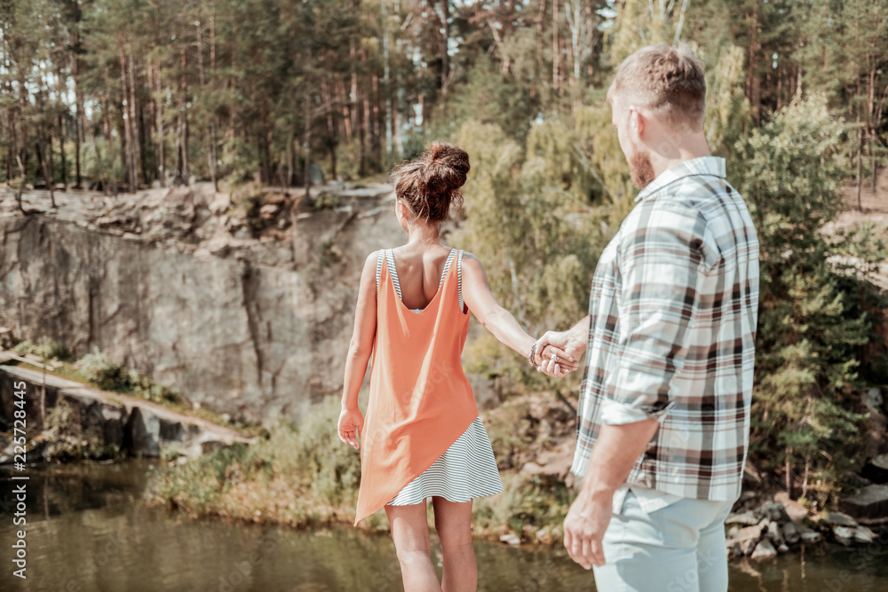 Holding hand. Bearded man holding hand of his dark-haired stylish girlfriend standing on the edge of rock near lake Holding hand. Bearded man holding hand of his dark-haired stylish girlfriend standing on the edge of rock near lake