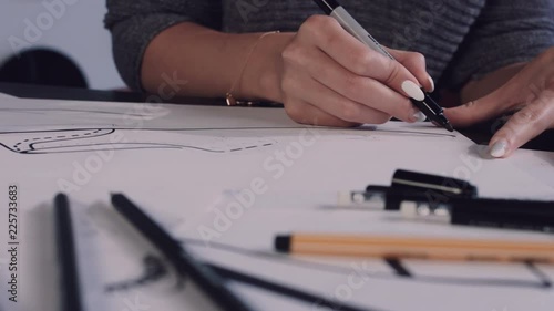 Attractive young woman sitting at the table in office, holding pencil and drawing sketch of shoes on paper. Shoes designer. 4K photo