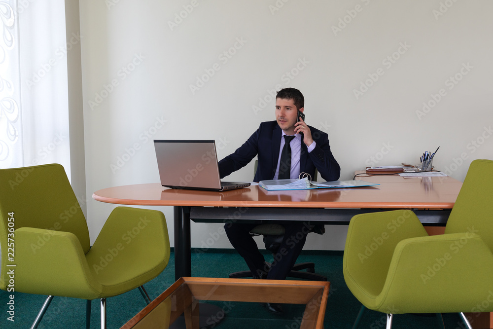 Young businessman wearing suit sitting alone at office desk talking on phone and working with laptop. Young businessman wearing suit sitting alone at office desk talking on phone and working with laptop.