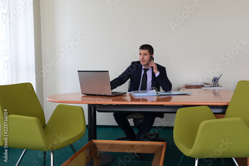 Young businessman wearing suit sitting alone at office desk talking on phone and working with laptop. 
