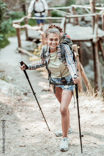 Beaming woman. Beaming curly dark-haired woman with backpack on her back feeling amazing while hiking with walking poles