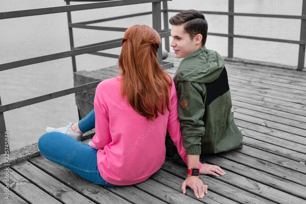 Boy and girl. Handsome young man wearing warm clothes while sitting on the pier with his pretty girlfriend and talking to her
