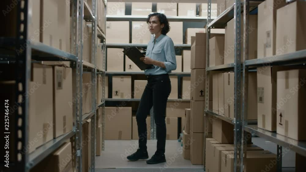 Female Inventory Manager Checks Stock, Writing in the Clipboard. Beautiful Woman Working in a Warehouse Storeroom with Rows of Shelves Full Of Cardboard Boxes, Parcels, Packages Ready for Shipment. Female Inventory Manager Checks Stock, Writing in the Clipboard. Beautiful Woman Working in a Warehouse Storeroom with Rows of Shelves Full Of Cardboard Boxes, Parcels, Packages Ready for Shipment.