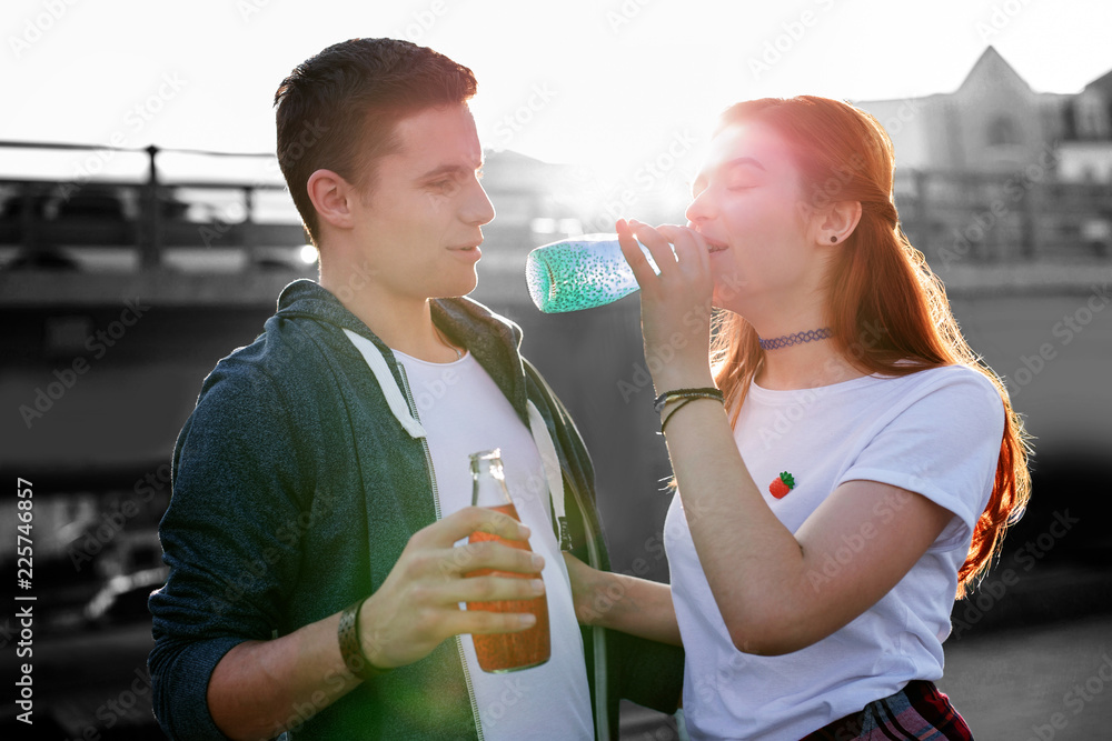 Drinking. Calm young girl standing next to her curious boyfriend and closing her eyes while drinking colorful cocktail Drinking. Calm young girl standing next to her curious boyfriend and closing her eyes while drinking colorful cocktail