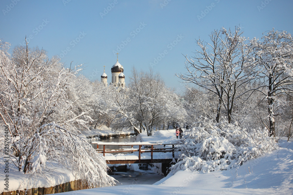 View of snow-covered river running in park with trees and bushes covered with snow, bridge and dome of church on background